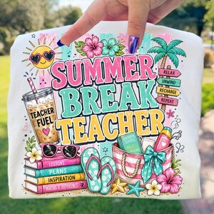 May include: A white t-shirt featuring a colorful graphic with the text SUMMER BREAK TEACHER. The design includes a sun with heart sunglasses, an iced coffee labeled TEACHER FUEL, a stack of books, flip-flops, a beach bag, and palm tree signs.