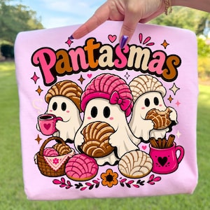 May include: A light pink t-shirt featuring a graphic of three white ghosts with concha bread, a pink mug with a heart, a basket of conchas, and cinnamon sticks in a pink mug. The text "Pantasmas" is printed in large, colorful, bubble-style letters.