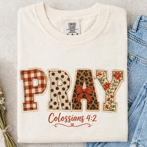 Może przedstawiać: Kremowy t-shirt z napisem PRAY zapisanym dużymi literami, z których każda ma inny wzór: czerwona kratka, czarne kropki, panterka z małą pomarańczową kokardką oraz wzór w kwiaty. Poniżej znajduje się nadruk z tekstem Colossians 4:2.