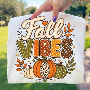 Puede incluir: Una camiseta blanca con un estampado gráfico que incluye el texto Fall VIBES en una fuente estilizada. El diseño presenta tres calabazas en naranja, verde y crema, con patrones de leopardo y lunares, rodeadas de hojas naranjas y verdes.