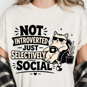 May include: A cream-coloured t-shirt featuring a black graphic of a cat wearing sunglasses, sitting in a chair, holding a mug with a heart, and a potted plant. The text reads: NOT INTROVERTED JUST SELECTIVELY SOCIAL in bold black lettering.