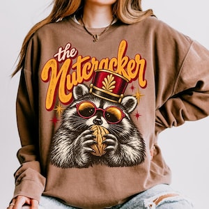 Könnte beinhalten: Ein braunes Sweatshirt mit Rundhalsausschnitt und einem Grafikdruck eines Waschbären, der eine rot-goldene Tambourmajor-Mütze und eine runde Brille mit rotem Rand trägt. Der Waschbär hält eine Walnuss, darüber steht "the Nutcracker" in Gelb.