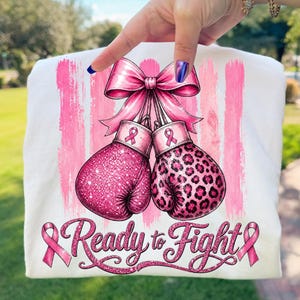 May include: A white t-shirt featuring a graphic of two pink boxing gloves tied together with a large pink bow. One glove has a glittery texture, the other has a leopard print. The text Ready to Fight is printed below with pink ribbons.