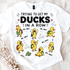 May include: A white t-shirt featuring a graphic of six yellow ducklings wearing backpacks, with the text "TRYING TO GET MY DUCKS IN A ROW" and "MRS. JONES!" repeated around the characters. The design includes small hearts, stars, and a pencil.