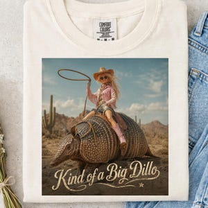 May include: Cream-colored t-shirt featuring a graphic of a doll dressed as a cowgirl riding an armadillo in a desert landscape. The text "Kind of a Big Dillo" is below the image. The shirt has a crew neck and short sleeves.