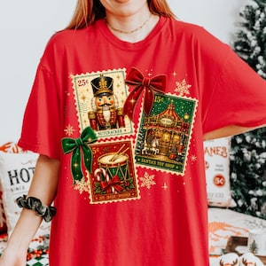 Könnte beinhalten: Ein rotes T-Shirt mit drei Briefmarken-Grafiken im Vintage-Stil. Die Briefmarken zeigen einen Nussknacker, einen Spielzeugladen und eine Trommel. Das Design umfasst eine große rote Schleife, eine grüne Schleife und goldene Schneeflocken. Der Text lautet 25c, 15c, 5c, NUTCRACKER, SANTA'S TOY SHOP und JOY MAGIC WONDER.