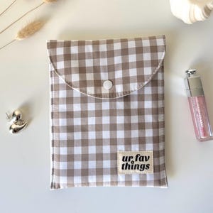 May include: A beige and white gingham pouch with a snap closure. The pouch features a label that reads "ur. fav things". A pink lip gloss and a silver duck-shaped object are also visible.