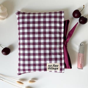 May include: A rectangular pouch featuring a dark purple and white gingham pattern with a fabric label that reads ur fav things. The pouch includes a dark purple ribbon tie on the side, providing a secure closure for the soft, structured textile item.