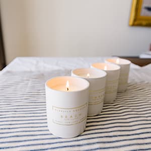 May include: Four white cylindrical candles with gold lettering and lit wicks are arranged on a blue and white striped surface. The front candle reads "Beach" and "Coconut Palm Leaves". The candles are part of a home decor collection.