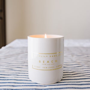 May include: A lit white candle in a cylindrical container with gold lettering that reads "Bluelily Candles BEACH 100% soy wax". The candle is on a blue and white striped surface. The candle's scent includes coconut, palm leaves, and almond.