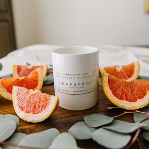 May include: A white candle with gold lettering that reads "Bluelily Candles Grapefruit 100% Soy Wax Grapefruit Mangosteen Zest" sits on a wooden surface. Slices of grapefruit and eucalyptus leaves surround the candle, creating a fresh, natural aesthetic.