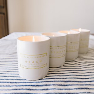 May include: Four white candles with gold lettering and trim are lit, sitting on a blue and white striped tablecloth. The candles have labels that read "Beach" and "Summer". The candles are cylindrical and have a clean, modern aesthetic.