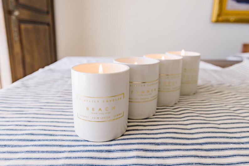 May include: Four white candles with gold lettering and trim are lit, sitting on a blue and white striped tablecloth. The candles have labels that read "Beach" and "Summer". The candles are cylindrical and have a clean, modern aesthetic.