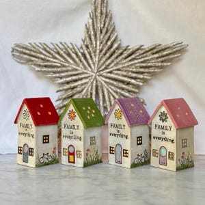 May include: Four decorative miniature houses with red, green, purple, and pink roofs. Each house has a white facade with a door, windows, and the text "FAMILY is everything." The houses are decorated with flowers and a bicycle. A large, glittery star is in the background.