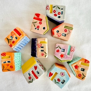 May include: A collection of colorful ceramic house-shaped ornaments. Each miniature house features unique designs, including red heart and floral accents, and varying roof and wall colors like orange, blue, yellow, and pink. The houses are arranged on a white surface.