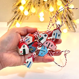 May include: A collection of small, hand-painted ceramic house ornaments in various colors including red, blue, pink, and white. Each miniature house features a painted door, windows, and floral accents, suspended by red and white twisted twine.