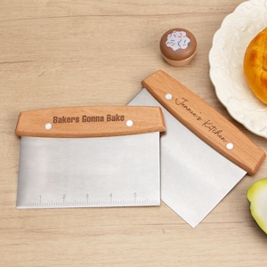 Puede incluir: Dos cortadores de masa con mangos de madera y cuchillas de acero inoxidable. Los mangos están grabados con "Bakers Gonna Bake" y "Jennie's Kitchen".