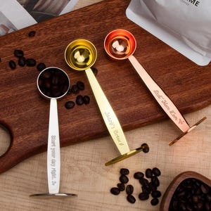 Könnte beinhalten: Drei goldfarbene Messlöffel mit gravierten Sprüchen. Die Löffel liegen auf einer Holzoberfläche mit Kaffeebohnen, die um sie herum verstreut sind. Die Sprüche lauten "Start your day right", "Just add's Brew" und "Eat, Drink, Love, Sip".
