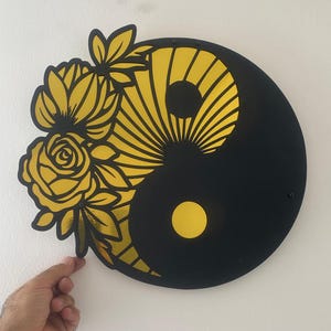 Könnte beinhalten: Ein schwarzes und gelbes Yin-und-Yang-Wandkunstwerk. Das Design zeigt ein Blumenarrangement auf der gelben Seite. Die Blumen sind gelb mit schwarzen Umrissen. Das Werk ist an einer weißen Wand befestigt.