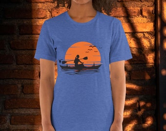 Puede incluir: Una camiseta azul jaspeada con un gráfico de una persona en kayak frente a una puesta de sol. El gráfico es de color naranja y negro.