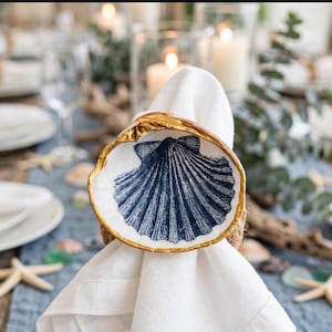 May include: A white fabric napkin held by a decorative napkin ring featuring a natural seashell. The interior of the shell is painted with a dark blue fan-shaped scallop design, and the outer rim of the shell is finished with a metallic gold trim.