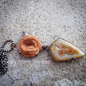 Baryte Rose sits above a shield shaped ocean jasper in cream with distinct orbs of orange accented by green.