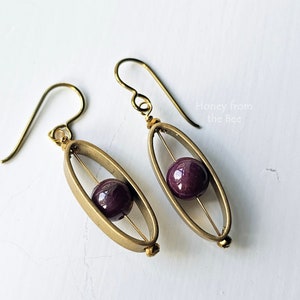 Peut inclure: Boucles d'oreilles ovales en or avec une perle violette suspendue à l'intérieur de chaque ovale.
