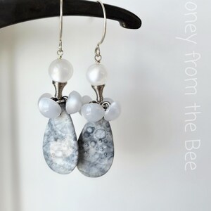 Puede incluir: Un par de pendientes colgantes con ganchos plateados. Cada pendiente presenta una cuenta redonda blanca, un grupo de pequeñas cuentas azul claro y una piedra en forma de lágrima, gris y blanca. Los pendientes cuelgan de un objeto negro curvado.