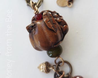 May include: A necklace featuring a brown carved pumpkin pendant with a small mouse, a red bead, a green bead, a metallic acorn charm, and a faceted red bead. The text Honey from the Bee is visible on the left side of the image. The chain is bronze.