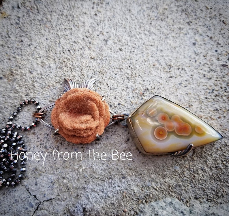 Baryte Rose sits above a shield shaped ocean jasper in cream with distinct orbs of orange accented by green.