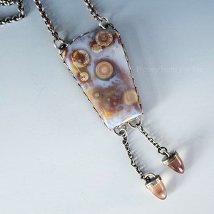 May include: A silver necklace with a large, white, brown, and beige agate pendant with a swirling pattern. The pendant is framed in silver and hangs from a silver chain with two silver tassels.