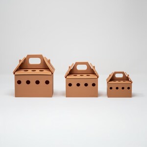 May include: Three terracotta-colored boxes with a house-like design. Each box has a handle on top and a series of circular holes on the front. The boxes are of varying sizes, with the largest on the left and the smallest on the right.