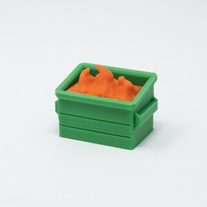May include: A miniature green dumpster with orange contents, possibly representing flames or waste. The small container has a rectangular shape with horizontal lines and a handle on each side. The contents are a bright orange color.