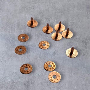 May include: A collection of copper-coloured rivets and washers on a grey surface. The rivets have a pointed top and a circular base. The washers feature a central hole and Japanese characters. These fasteners are suitable for leatherwork or similar crafts.