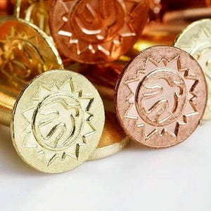 Pode incluir: Close-up de várias moedas metálicas em tons de ouro, cobre e bronze. Cada moeda apresenta um desenho de sol em torno de um emblema central. As moedas estão empilhadas, criando um efeito texturizado e em camadas.