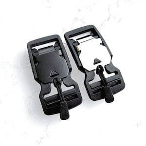 May include: Two black plastic buckles with a metal insert, each featuring a cord loop. The buckles are designed with a secure locking mechanism. The image is taken from a top-down perspective against a white background.