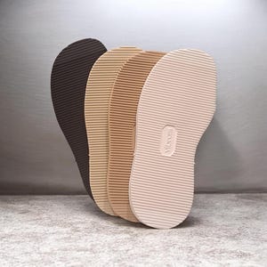 May include: A set of four shoe soles arranged in a fan shape, featuring a dark brown, a tan, a light brown, and a cream-coloured sole. Each sole has a horizontal ribbed texture and the Vibram brand name embossed in the centre of the cream sole.