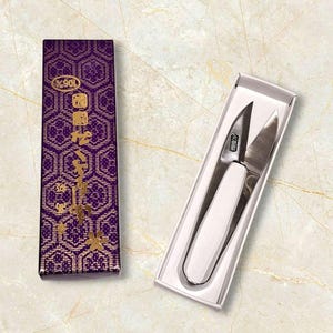 May include: Silver-bladed scissors with a white handle, displayed in a white box. Next to it, a purple box with gold text and a geometric design. These scissors are likely for crafts or sewing.