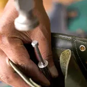 K&ouml;nnte beinhalten: Eine Person, die einen Hammer verwendet, um einen Metallniet in einen braunen Lederstiefel zu befestigen.