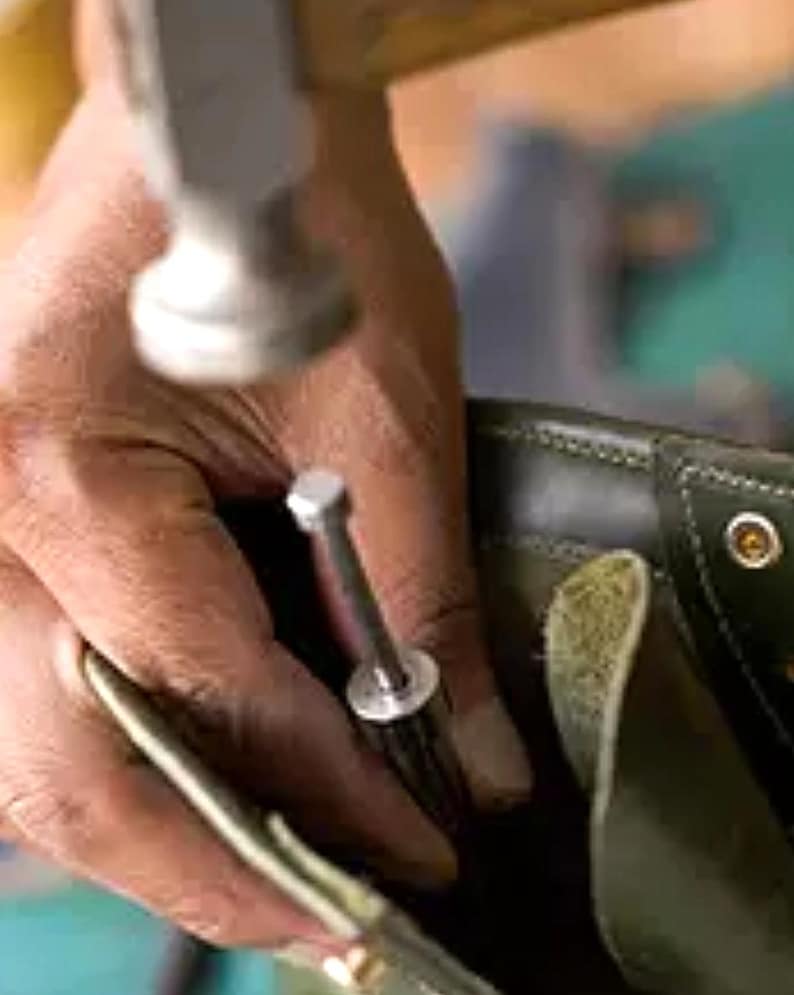 K&ouml;nnte beinhalten: Eine Person, die einen Hammer verwendet, um einen Metallniet in einen braunen Lederstiefel zu befestigen.