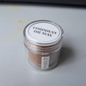 Könnte beinhalten: Ein kleines, klares Glas mit braunem Wachs darin. Der Deckel hat ein weißes Etikett mit der Aufschrift "Cordovan Oil Wax".