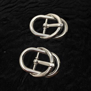Pode incluir: Duas fivelas de cinto prateadas e brilhantes. Cada fivela tem formato oval com uma barra central e um design de laço. As fivelas são feitas de metal refletivo e estão sobre um fundo escuro e texturizado.