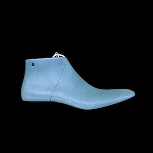 Può includere: Una forma per scarpe azzurra, utilizzata nella fabbricazione di calzature, è mostrata su sfondo nero. La forma ha una punta affilata e un piccolo foro vicino al tallone. Un cordino bianco è attaccato in alto.