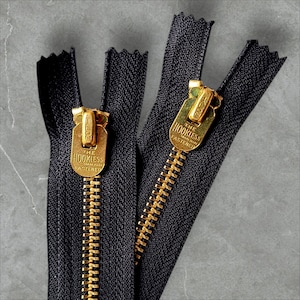 May include: Two black zipper tapes with gold metal teeth and pull tabs. The pull tabs have the text "The Hookless Fastener" on them.