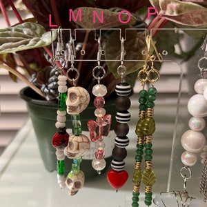 Puede incluir: Cinco pares de pendientes colgantes con diferentes dijes. Los dijes incluyen una calavera, una mariposa, un corazón y hojas. Los pendientes se muestran en un soporte blanco con las letras L, M, N, O y P.
