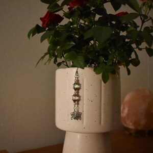 May include: A white ceramic planter with a rose bush and a decorative charm. The charm has pink beads, a silver-colored butterfly, and other decorative elements. The planter is on a wooden surface.