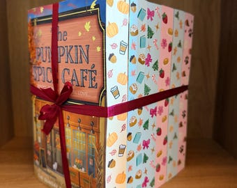 Puede incluir: Una pila de libros atados con una cinta de color rojo oscuro. La portada del libro frontal presenta una ilustración de una cafetería con el texto The Pumpkin Spice Cafe. Los lomos de los libros están decorados con patrones coloridos de calabazas, hojas, tazas de café y lazos.