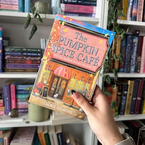 K&ouml;nnte beinhalten: Ein Buchcover mit einer Illustration eines Caf&eacute;s mit einem Schild, auf dem steht "The Pumpkin Spice Cafe It's the season to fall in love." Das Buch tr&auml;gt den Titel "The Pumpkin Spice Cafe" von Laurie Gilmore.