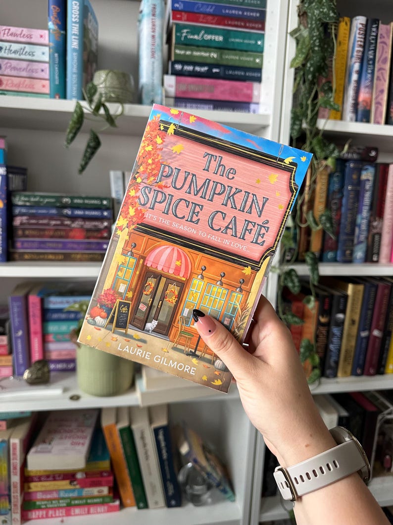 K&ouml;nnte beinhalten: Ein Buchcover mit einer Illustration eines Caf&eacute;s mit einem Schild, auf dem steht "The Pumpkin Spice Cafe It's the season to fall in love." Das Buch tr&auml;gt den Titel "The Pumpkin Spice Cafe" von Laurie Gilmore.