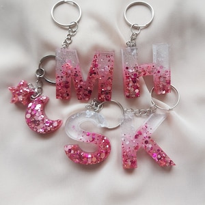 May include: Pink glitter resin letter charms with silver keyrings. The letters spell out "MSK" and there is a pink glitter star and crescent moon charm.